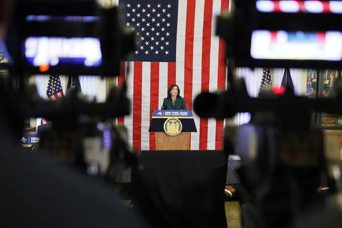 Gov. Kathy Hochul delivers a speech in front of an American flag as cameras record her.